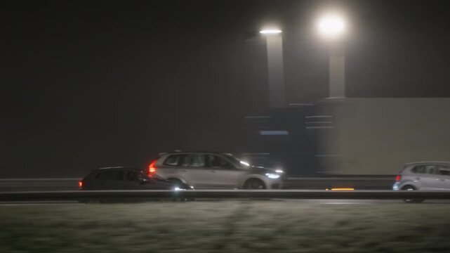 Side view of cars driving on a highway at night in thick, atmospheric fog. Streetlights glow, illuminating the poor visibility. A tracking shot follows vehicles as they travel on the road