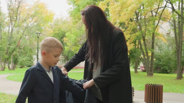 Park morning parent helps boy adjust jacket before school, gentle sendoff on bench under autumn trees, caregiver fastening coat, warm supportive interaction, calm preparation for school day
