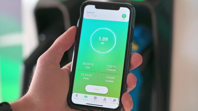 Close-up of hand holding smartphone with green charging app, electric vehicle charging in background.
