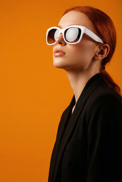 Stylish young redhead woman wearing white sunglasses and a black blazer posing against a vibrant orange background.