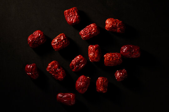 Dried red dates or Chinese jujube scattered on black background. Moody food photography with dramatic lighting, highlighting texture and vibrant red color.