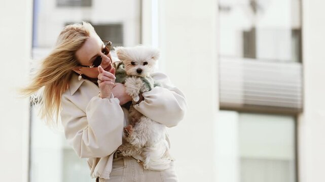 4K slow motion horizontal video of a fashionable woman with sunglasses holding cute mini Maltese dog in windy city street. Trendy urban lifestyle, movement, stylish pet owner concept.