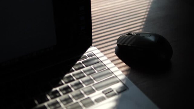 Cinematic close-up of wireless computer mouse and laptop keyboard under dramatic window blind shadows for late-night work concept