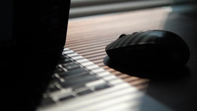 Cinematic close-up of wireless computer mouse and laptop keyboard under dramatic window blind shadows for late-night work concept