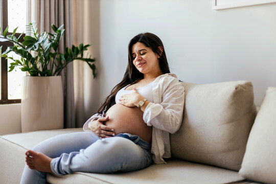 Pregnant woman sitting on a sofa touching her baby bump and relaxing, enjoying a calm and peaceful moment at home during pregnancy.