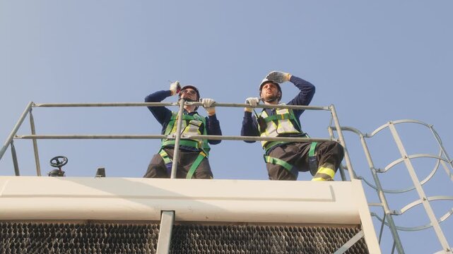Two industrial engineers in safety helmets and harnesses standing on a rooftop mechanical platform at a large industrial facility with solar panels.