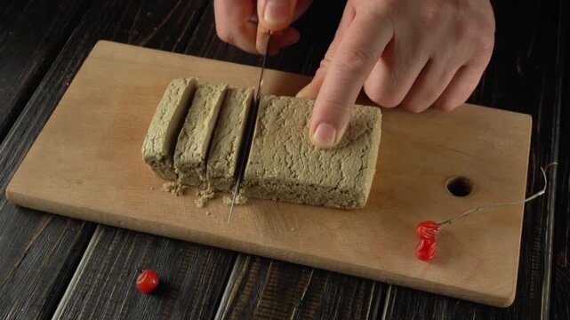 A hand with a knife cuts a block of halva on a wooden cutting board decorated with a red cherry, showing a clean kitchen environment with natural light