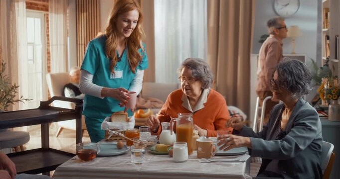 Nurse bringing food to elderly residents at dining table in assisted living facility. Seniors sharing breakfast and interacting in cozy home environment with care and support.