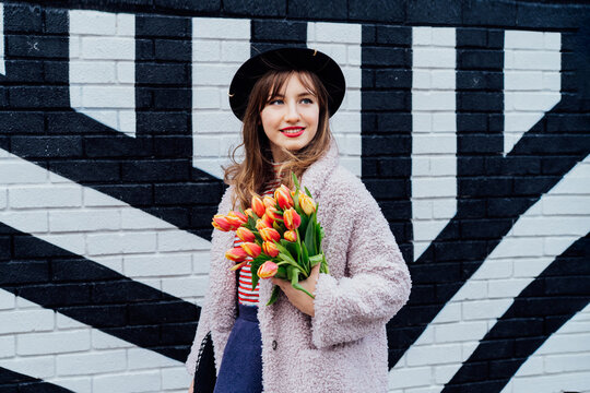 Portrait of happy. smiling young fashion woman holding bouquet of fresh tulip flowers on the painted brick wall background. Urban city street fashion. Spring mood. Happiness and positive vibes.
