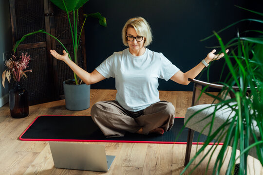 Middle aged woman practicing online yoga workout using laptop in modern Scandi dark interior home. Healthy Lifestyle. Sport activity for mature wellbeing. Fitness online training. Mindful movement