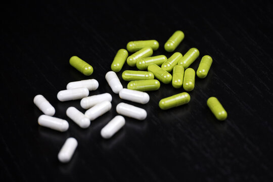Pills in white and green capsules scattered on a black wooden table. Background for herbal medicine, antibiotics, vitamins, dietary supplements