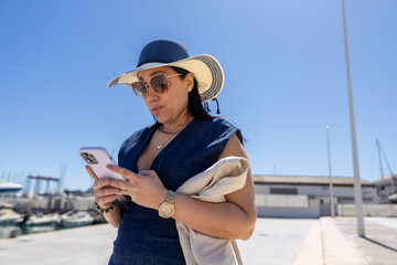 Fototapeta premium Woman in sun hat using smartphone at a marina on a sunny day.