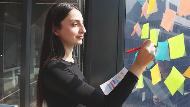 Young professional organizing ideas on colorful sticky notes while reviewing business chart near office window during planning session.