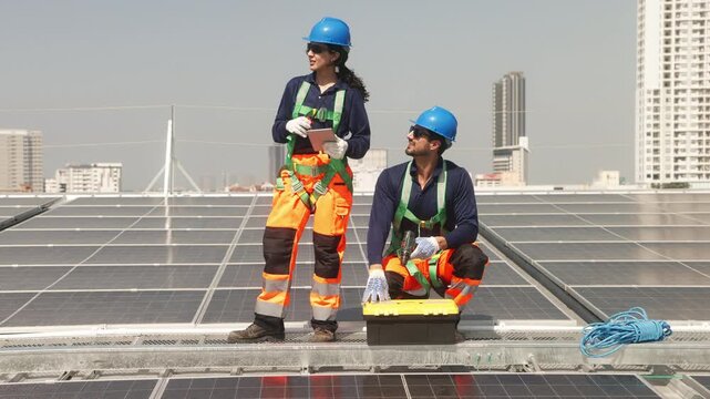 Two solar technicians working on rooftop photovoltaic installation, one drilling panel mounts while colleague checks system data on a tablet.