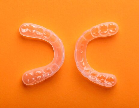 Two clear, molded plastic dental aligners sit on a vibrant orange background. Each curved piece mimics the shape of a dental arch, showcasing individual tooth impressions