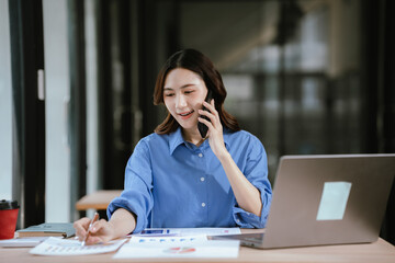 Obraz premium A young businesswoman talking on the phone while writing on financial charts at a desk with a laptop and coffee in a modern office.