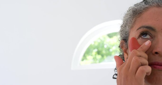 Senior African-American woman lifting hand, placing under-eye patch for care by window, copy space