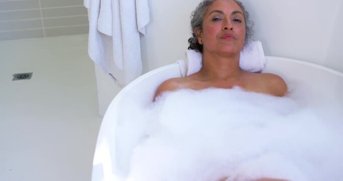 Senior African woman resting in bathtub, foam, towel headrest, camera pulling back revealing face