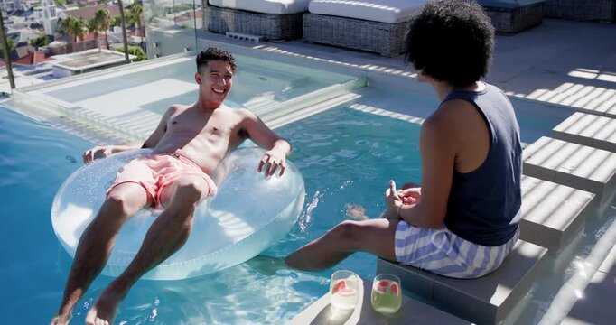 Two men in swimwear chatting after gesture, sharing drink, reclining on clear float at rooftop pool