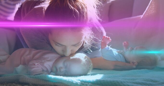 Kissing mother leaning over sleeping infant girls on couch, showing pink sleeper blue onesie