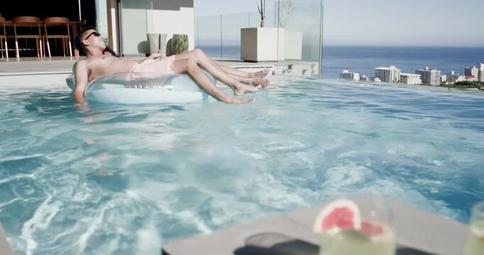 Camera pulling back, African American men relaxing in rooftop pool on float, pointing at city