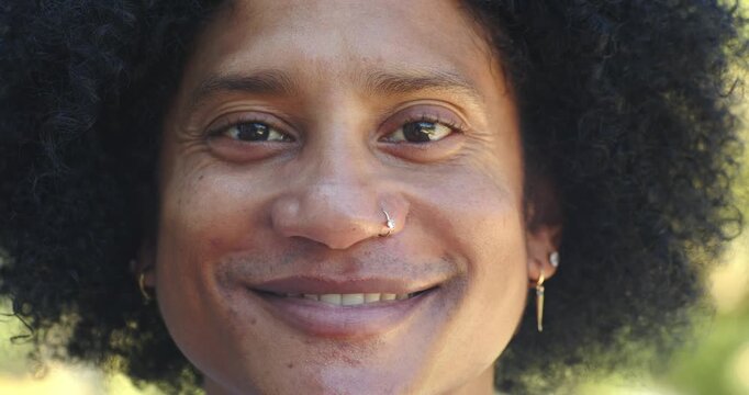African man smiling into open-mouthed laugh in sunlit park, showing nose hoop and multiple earrings