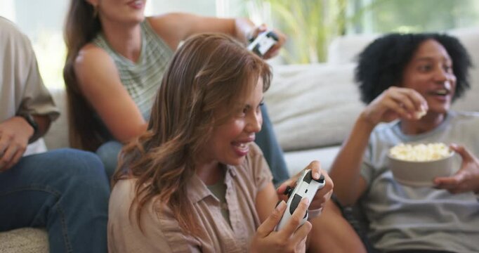 Diverse friends sitting on living room couch playing group game holding white controllers, popcorn