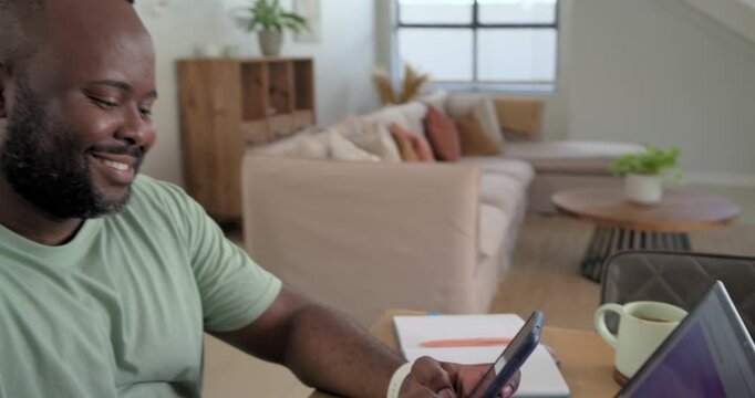African male seeing message on smartphone, typing reply on laptop, smiling in home office