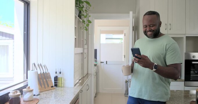 Vertical video: African American man checking phone text, smiling and sipping mug in kitchen