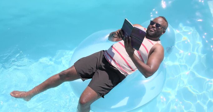 African American man reclining on blue float in pool reading book turning pages kicking relaxing