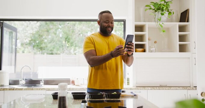Phone pinging African American man tapping phone laughing, glancing at meal boxes on kitchen island