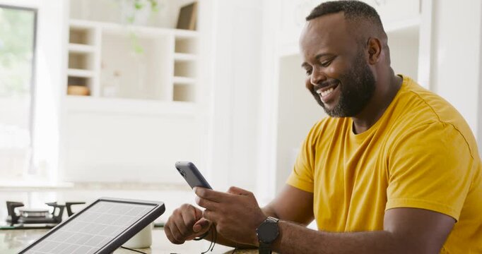 30s African man picking phone adjusting cable checking solar charge at kitchen counter mustard tee
