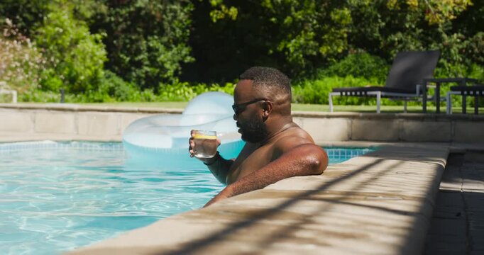 Mid adult African American man raising glass with lemon, sipping while seeking relief by pool float