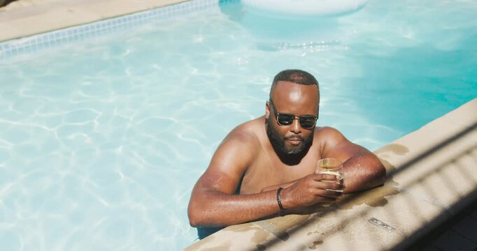 African man shirtless leaning at pool edge, sipping from glass in sun, gazing at camera