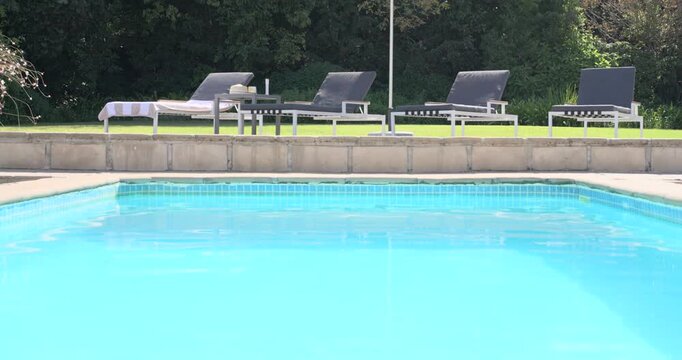Tiled pool rippling under sun in backyard while frame shifting up to show umbrella, loungers, towel