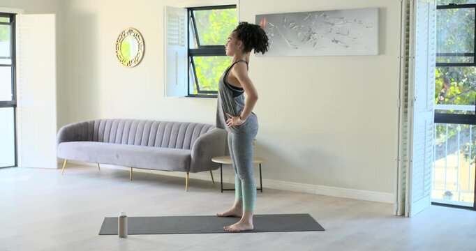 African American woman standing on yoga mat, reaching twisting folding for stretch at home