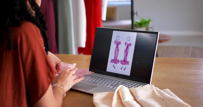 Referencing laptop sketch, woman sitting at worktable holding sheer pink fabric to match gowns