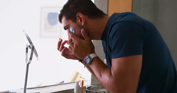 Leaning toward magnifier at vanity, man examining applying under-eye patches pressing to hydrate