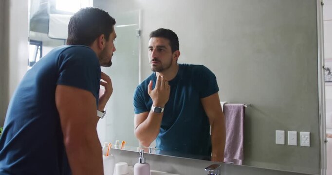 Adult male entering home bathroom and leaning at sink, examining chin in mirror, checking stubble