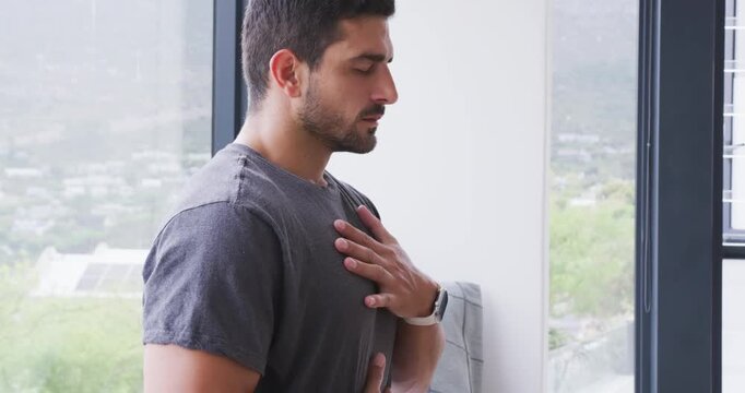 Man closing eyes, placing hands on chest and abdomen, focusing on breath beside window bench, mat