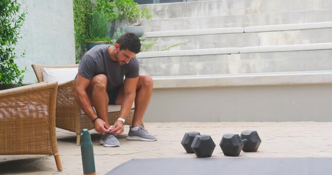 Man mid 20s tying and adjusting shoelaces on patio bench preparing for workout with dumbbells