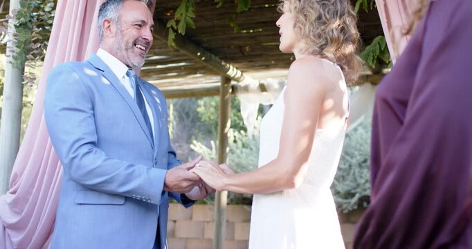 Couple holding hands pink-drape pergola groom speaking vows gowned bride listening Indian officiant