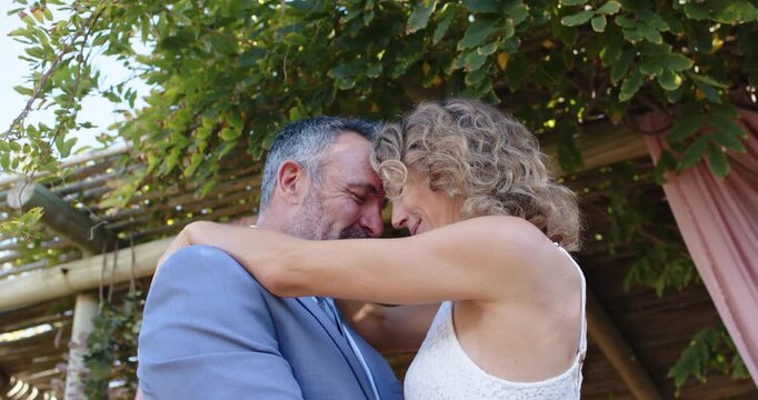Couple pressing foreheads, holding each other under pergola, sharing affection in white lace dress