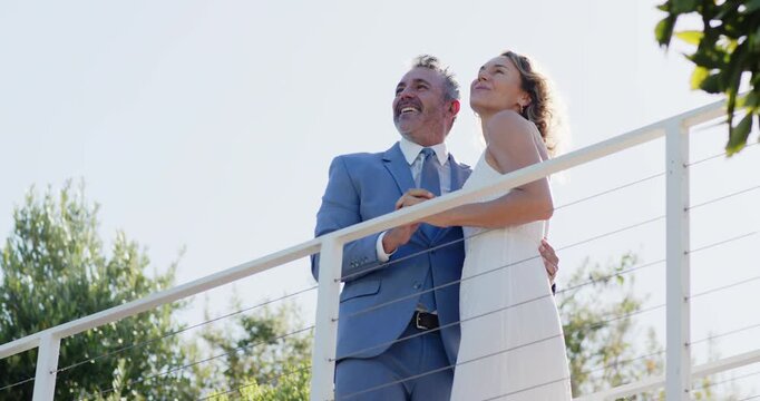 Bride and groom in formal wear, groom taking bride's hand embracing gazing skyward on white railing