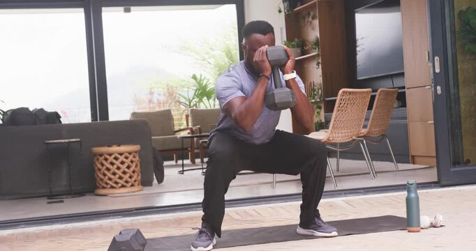 African man in sportswear gripping hex dumbbell, doing squats, lunges at home mat for strength