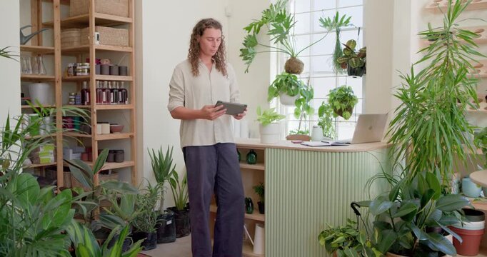 Nonbinary adult using tablet, glancing at laptop, smiling, nodding at offscreen voice in plant shop