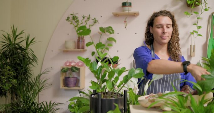Nonbinary florist in plant shop in apron lifting plants, placing in black stand styling display