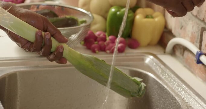 Adult African man bringing leeks, rinsing under running faucet into colander, trimming for cooking