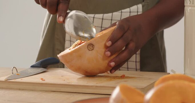 Man wearing apron scraping butternut squash seeds with spoon on cutting board at kitchen counter