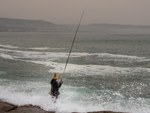 Fisherman standing on rocky shoreline casting line into rough Atlantic waves at Anchor Point near Taghazout, Morocco
